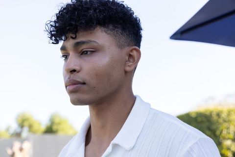African american man standing in backyard under umbrella wearing white knit polo, pensive