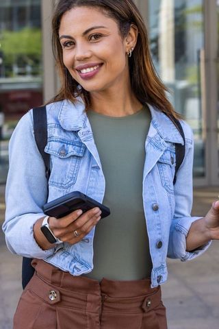 African american woman smiling and holding smartphone, wearing denim jacket outside glass building