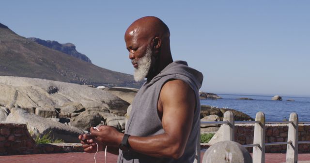 Senior man with a gray beard adjusting earbuds by the seaside, preparing for an exercise routine. Ideal for content related to active lifestyles, health and fitness, technology use in outdoor activities, or scenes depicting natural beauty and wellness.