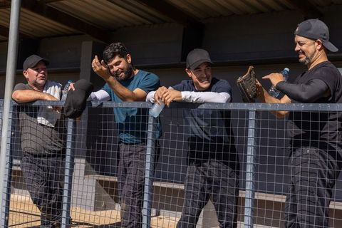 Baseball teammates laughing and leaning on dugout fence holding caps and gloves with water bottles