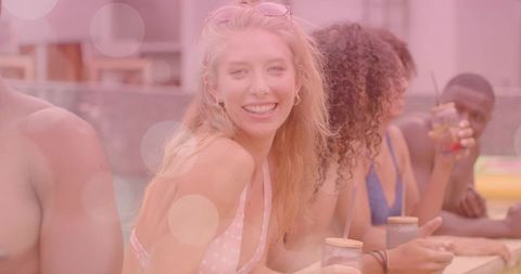 Smiling blonde woman in pink polka-dot bikini leaning on pool deck with mason jar drink