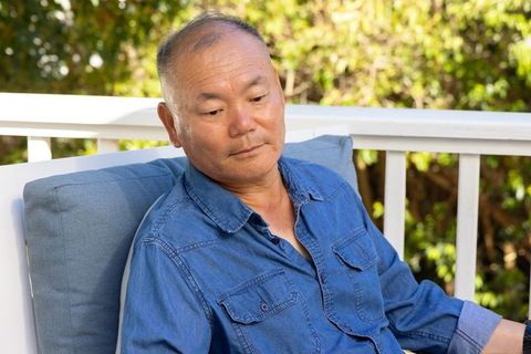 Mature asian man relaxing on sunlit porch in denim shirt contemplative casual lifestyle