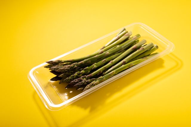 Fresh asparagus in a plastic container on a vibrant yellow background. Ideal for use in healthy eating campaigns, organic food promotions, diet and nutrition articles, and culinary blogs. The bright background adds a modern and fresh feel, perfect for advertisements and social media posts.