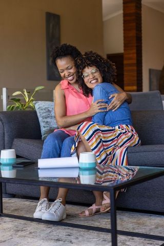 Laughing friends hugging on sofa sharing cozy moment with mugs and colorful skirt