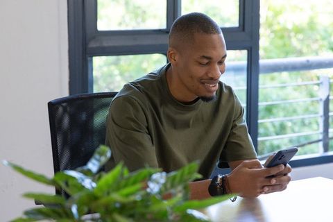 African american man smiling while using smartphone at sunlit home office desk