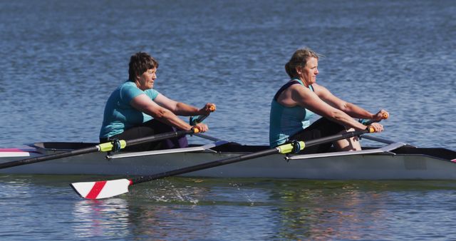 Two women rowing in unison on a calm lake, demonstrating teamwork and coordination. This image is perfect for promoting outdoor sports and fitness, emphasizing the importance of partnership, resilience, and synchronized effort. Ideal for use in health and wellness campaigns, sports event promotion, team building activities, and articles about fitness and active lifestyles.