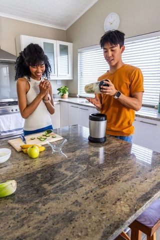 Young diverse couple making green smoothie at bright modern kitchen island for healthy breakfast
