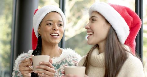 Two friends sharing cozy holiday coffee, wearing santa hats, smiling by window