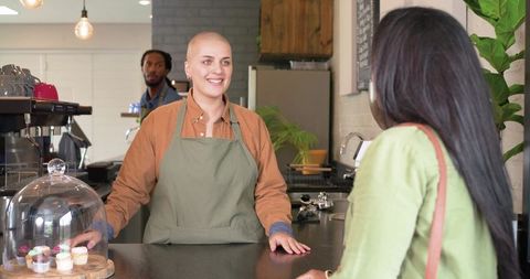 Smiling bald barista serving cupcakes to indian customer at modern cafe counter
