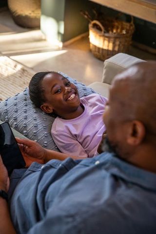 African american father bonding with daughter using tablet on sofa in sunlit living room