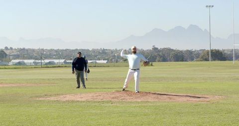 Pitcher delivering windup on sunlit mound, umpire watching, outfielder awaiting play