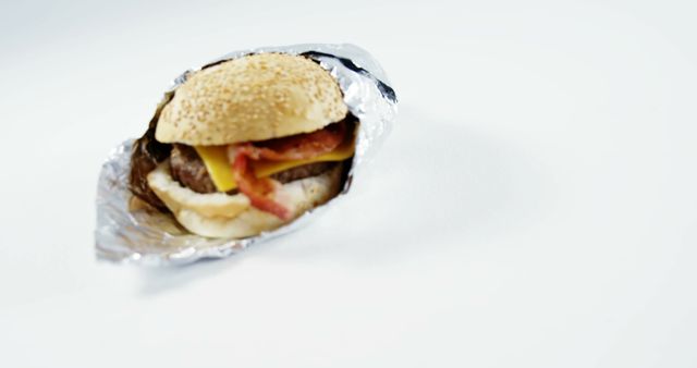 A bacon cheeseburger wrapped in foil sits against a white background, with copy space. Its presentation suggests a quick, convenient meal option, often associated with fast food.