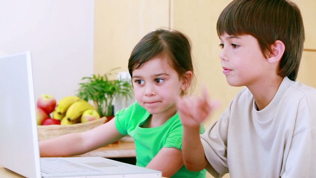 Two children work on a laptop in a bright kitchen, enjoying and having fun, making funny faces. Perfect for use in educational content, technology ads, family-related promotions, or child entertainment websites.