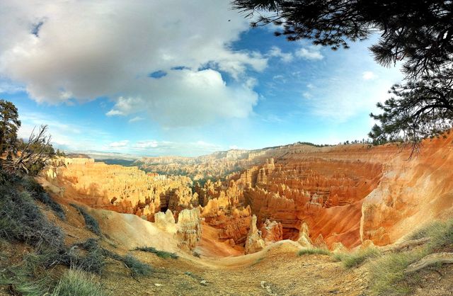 Panoramic view of Bryce Canyon National Park captures stunning red rock formations under a blue sky scattered with clouds. Ideal for travel brochures, nature blogs, educational material about geology, and posters for promoting adventure tourism.