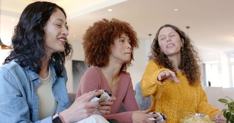 Diverse women playing video games on couch reaching for chips, cozy modern living room