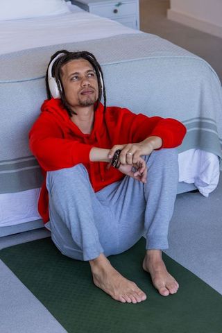 African american man listening to music and relaxing on yoga mat at foot of bed at home