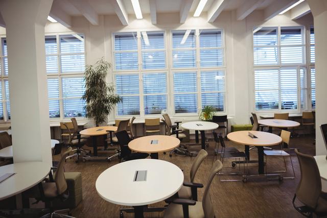 Modern empty office space featuring round tables and chairs arranged in an open plan layout. Large windows allow natural light to flood the room, creating a bright and inviting atmosphere. Ideal for illustrating contemporary work environments, corporate settings, and professional spaces. Suitable for use in business presentations, office design portfolios, and articles on modern workplace trends.