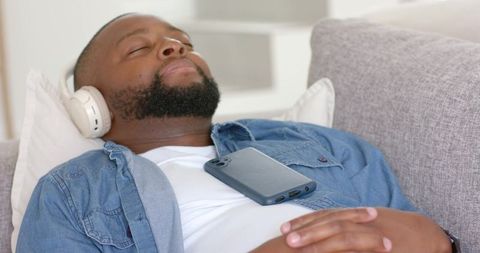 Relaxing man listening to music on couch with wireless headphones and phone on chest