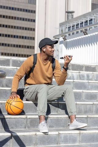 African american man sitting on city steps holding basketball using smartphone and earbuds
