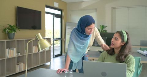 Diverse female coworkers wearing headscarves collaborating around laptop in modern office