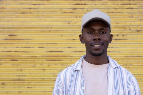 African american man standing against yellow shutter smiling in beige cap, striped shirt