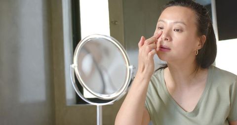 Asian woman applying under-eye skincare while looking into round tabletop mirror