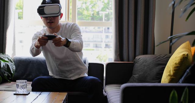 Asian man wearing VR headset, interacting with virtual environment in living room. Shows excitement, focus on enjoyment of advanced gaming technology. Use for technology, entertainment, virtual reality, and lifestyle concepts.