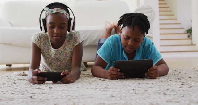 African american brother and sister using electronic devices lying on the floor at home. family, togetherness and happiness concept