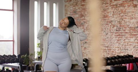 Asian woman stretching in gym pulling hoodie over light blue activewear near dumbbells