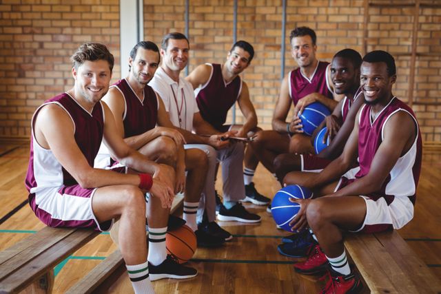 Group of basketball players and their coach sitting on a bench in an indoor court, smiling at the camera. They are wearing matching uniforms and holding basketballs. This image can be used for sports team promotions, coaching advertisements, teamwork concepts, and athletic training materials.