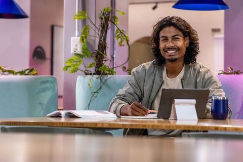 Smiling asian man using tablet and studying in modern coworking lounge with plants