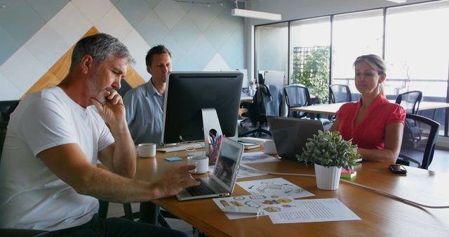 Professionals collaborating and having a discussion around a table in a modern office space with large windows. Ideal for business, teamwork, office decor, modern workspaces, collaborations, and professional settings. Use this to illustrate modern office environments, business decks, teamwork importance, and productive discussions.