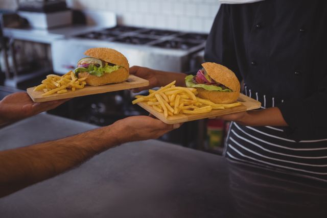 Waitress handing over two plates of burgers and fries to a coworker in a cafe kitchen. Ideal for use in articles about restaurant service, teamwork in hospitality, fast food, and culinary environments.