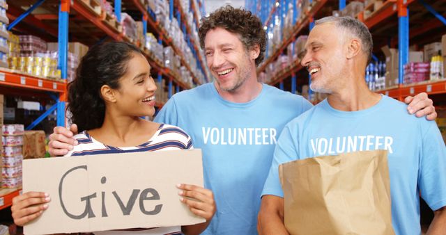 Three volunteers are smiling and holding a sign that says 'Give' in a warehouse stocked with supplies. Ideal for illustrating community involvement, charitable activities, teamwork, and humanitarian efforts in marketing materials, websites, and social media posts.