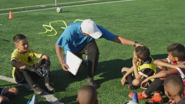 Biracial male soccer coach instructing a diverse group of young boys during a sunny outdoor training session. Perfect for use in content related to youth sports, teamwork, coaching, physical education programs, summer activities, and the importance of mentorship in sports.