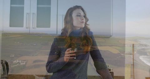 Woman gazing through kitchen window at coastal landscape while holding glass, contemplative