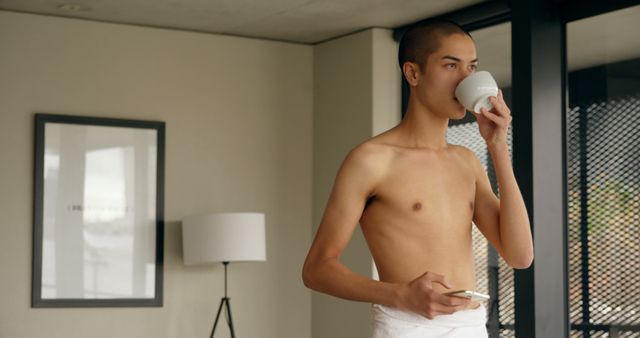 A young man is drinking coffee while using a smartphone, wrapped in a towel at home. Use this image for storylines related to morning routines, leisure at home, or modern lifestyles.