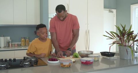 African american father teaching son chopping vegetables on modern kitchen island