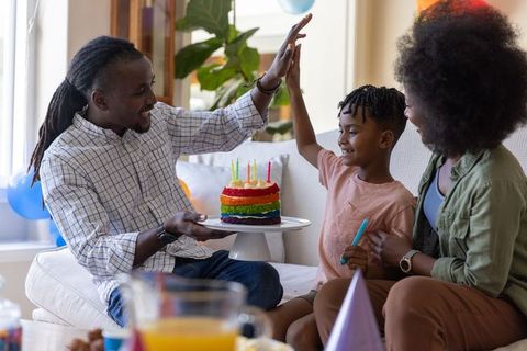African american family celebrating child birthday on couch with rainbow cake and balloons