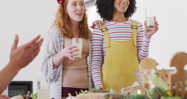 Two women standing with drinks, smiling and toasting at a casual gathering with friends. A mixed-race group engaged in friendly conversation and enjoying each other's company. Suitable for use in social event promotions, friendship-themed advertisements, or lifestyle blog articles highlighting joy and togetherness.