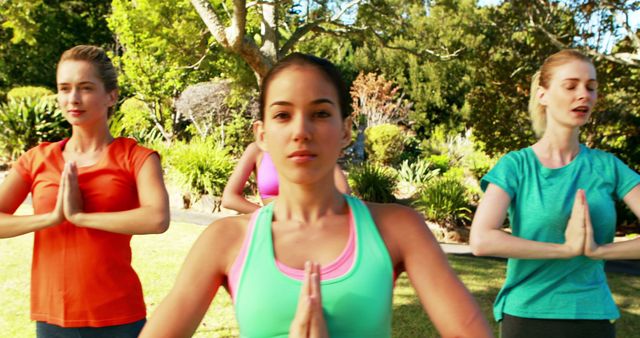 Women practicing yoga outdoors in a park with lush greenery and summer vibes. Ideal for use in promoting outdoor fitness classes, yoga retreat advertisements, and articles on the benefits of exercise and meditation.