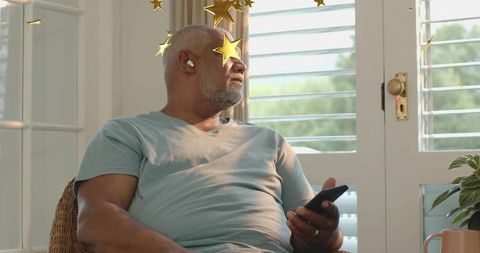 Mature man relaxing with earbuds and smartphone near sunlit shutters with gold star overlay