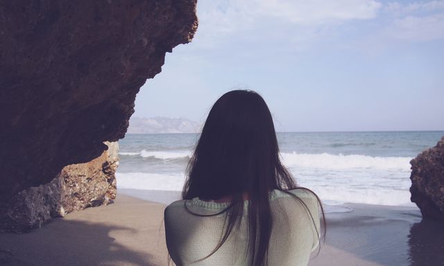 A woman with long hair facing ocean waves, standing between rocks on a scenic beach. Ideal in travel brochures, coastal escape advertisements, relaxation concepts, websites promoting natural beauty or mindfulness.