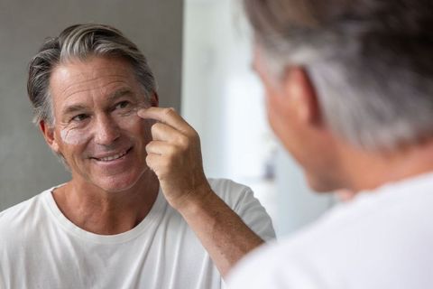Mature man applying under-eye patches at mirror smiling self-care grooming routine