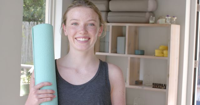 Young woman smiling while holding a rolled yoga mat in a wellness studio with yoga blocks and props in the background. Ideal for use in marketing materials for yoga classes, health and wellness programs, fitness studios, or articles promoting healthy lifestyles.