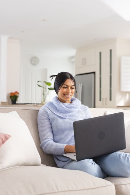 Middle Eastern woman sitting on a sofa, smiling while using a laptop. Ideal for illustrating remote work, modern lifestyle, home comfort, and technology use in everyday life. Perfect for blogs, articles, and advertisements related to working from home, technology, and lifestyle.