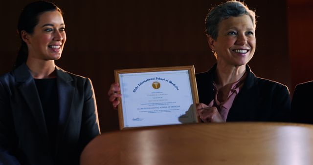 Woman receiving an award at a presentation ceremony while smiling proudly next to another woman, indicating the joy and happiness of achieving recognition. Ideal for use in articles, blogs, and promotions about professional achievements, motivation, and awards.