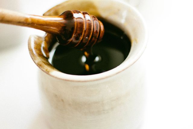 Close-up of honey being dripped from a wooden honey dipper into a ceramic pot. Ideal for use in articles and marketing materials related to food, organic produce, healthy eating, and natural ingredients. Perfect for recipe blogs, cooking websites, and nutrition-related content.