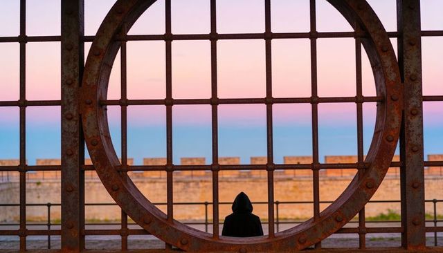 Solitary hooded figure framed by rusted circular iron grid over pastel seascape sunrise