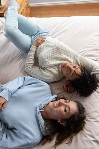 Two diverse women lounging on bed in cozy neutral bedroom showing ring and casual jewelry
